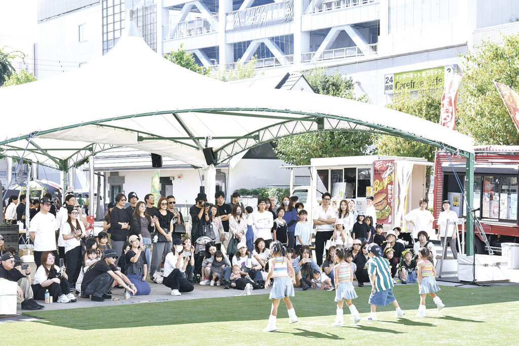 人工芝のステージで子どもたちがダンスを披露し、大型テントの下で来場者が見物した。公園の周りにはキッチンカーも並んだ